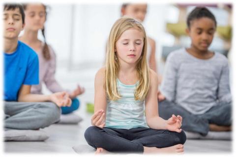 Blond girl sitting crossed leg on floor in yoga pose with children sitting behind her i similar pose.