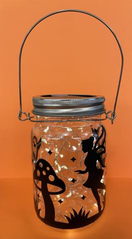 Orange background and a mason jar with a fairy, mushroom and grass silhouette. The lantern has a string of white lights inside. 