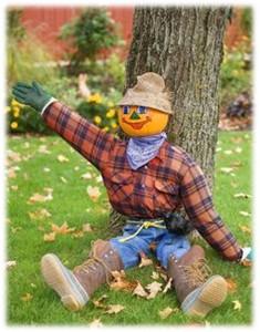 Scarecrow with a painted pumpkin face sitting on the ground propped up by  a tree. Scarecrow is wearing a flannel shirt, blue jeans and boots.