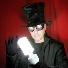 A man wearing a black magician's hat, black googles, black gloves, black jacket and black shirt holding a spiral shaped white light bulb with a red curtain in background. 