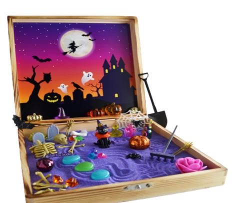 Wooden box filled with purple sand and Halloween objects (skeletons, stones, graves, rake, and spiders). Halloween background with a spooky house, pumpkin and tree. Two ghosts, bats, and a witch flying over the moon. 