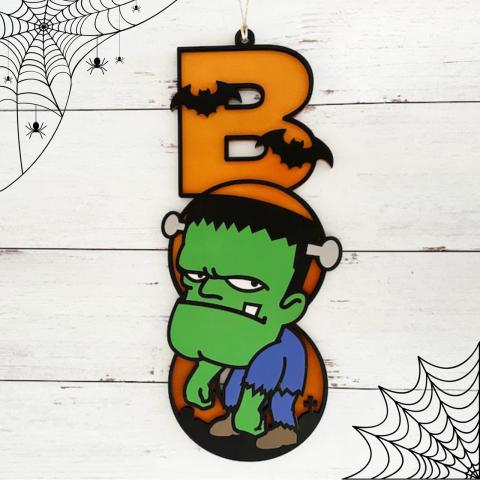 The letters BOO spelled with Orange "B" with two bats, Green Frankenstein Head for the letter "o", and Frankenstein's body for the second "O" on a white background with spider webs. 