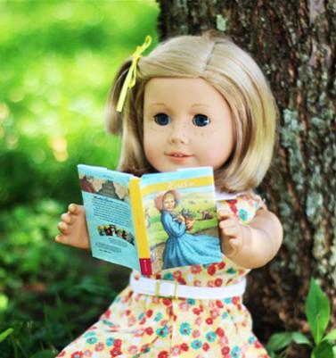 Doll with short blonde hair holding a book sitting against a tree trunk. 